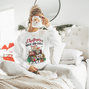 Smiling woman in a 'Christmas Movies & Chill' jumper, settled comfortably in a cozy home setting, ready to watch Christmas movies. The jumper, featuring retro televisions stacked in a Christmas tree shape with classic Christmas movies on their screens, adds a playful and nostalgic touch to the scene. 