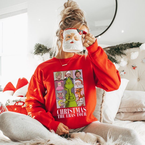 woman wearing the 'Festive Film Favourites' Christmas jumper, comfortably seated in a cozy home setting with a Christmas-themed mug in hand. The jumper, adorned with a grid of classic Christmas movies, adds a colourful and nostalgic touch. She's surrounded by festive decor, including a twinkling Christmas tree and warm, glowing lights, creating an inviting and festive atmosphere. The scene captures the essence of a relaxed, movie-themed Christmas evening.